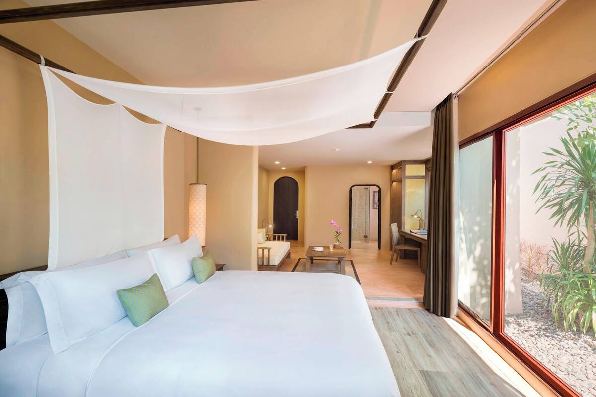 A spacious bedroom with a large bed, a small table, and a view of a garden through a large window.