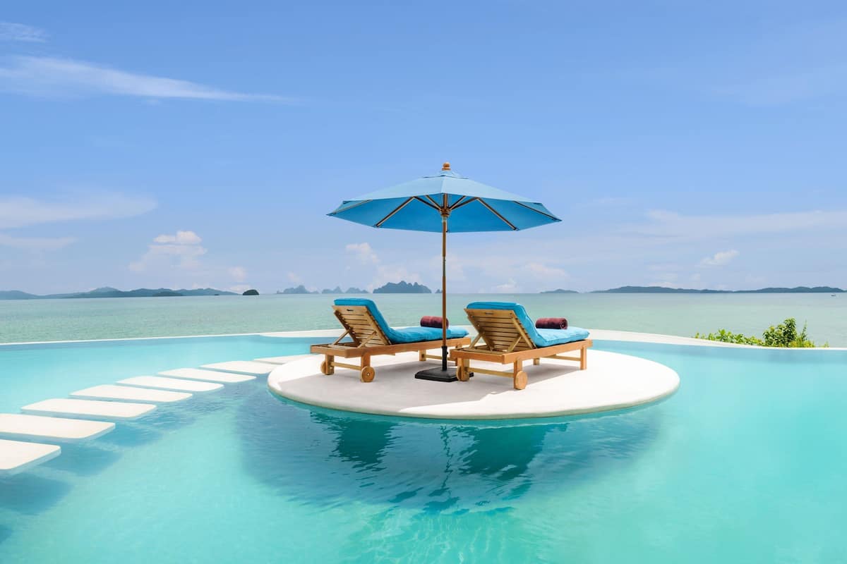 A poolside area with two lounge chairs and a blue umbrella, surrounded by clear blue water and a view of distant islands.