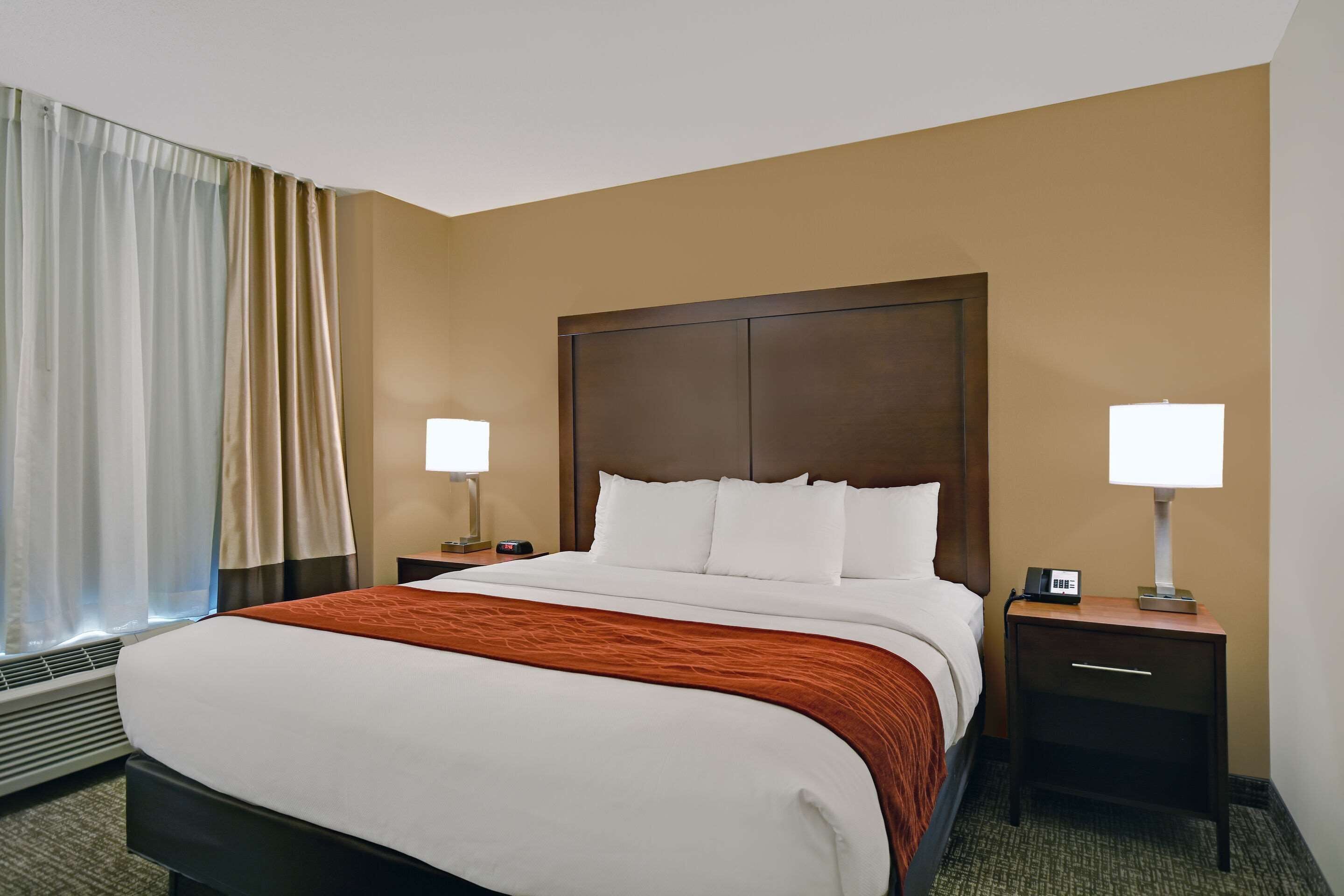 Habitación de hotel con una cama grande, dos mesitas de noche con lámparas, un ventanal con cortinas y un equipo de aire acondicionado instalado en la pared.