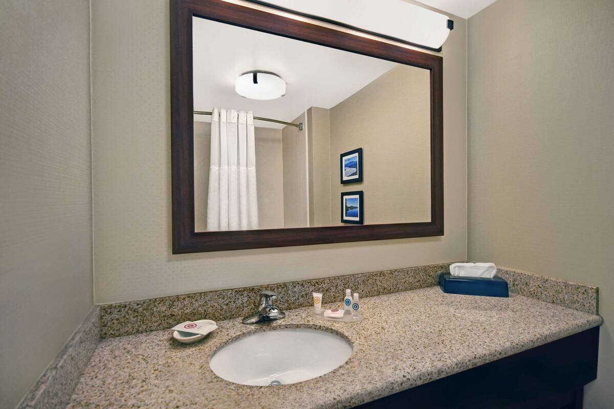 Une salle de bain avec un comptoir en granit, un lavabo blanc, un miroir avec un cadre en bois, un porte-serviettes et une petite étagère contenant des articles de toilette.
