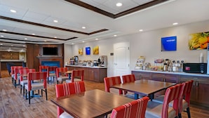 A hotel dining area with multiple tables and chairs, a kitchen area with a coffee station, and a TV mounted on the wall. - Comfort Inn & Suites Wilton (Wilton)