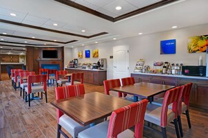 Un comedor de hotel con varias mesas y sillas, una zona de cocina con estación de café, y un televisor montado en la pared.