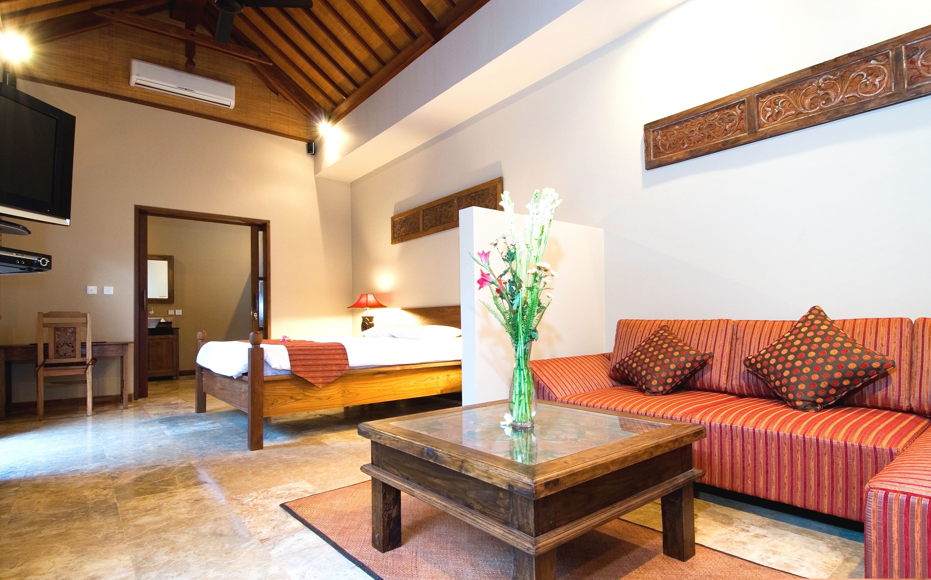 Ein Hotelzimmer mit Holzboden, einem Bett mit roter Decke, einer Couch mit gemusterten Kissen, einem Glastisch und einer Vase mit Blumen.
