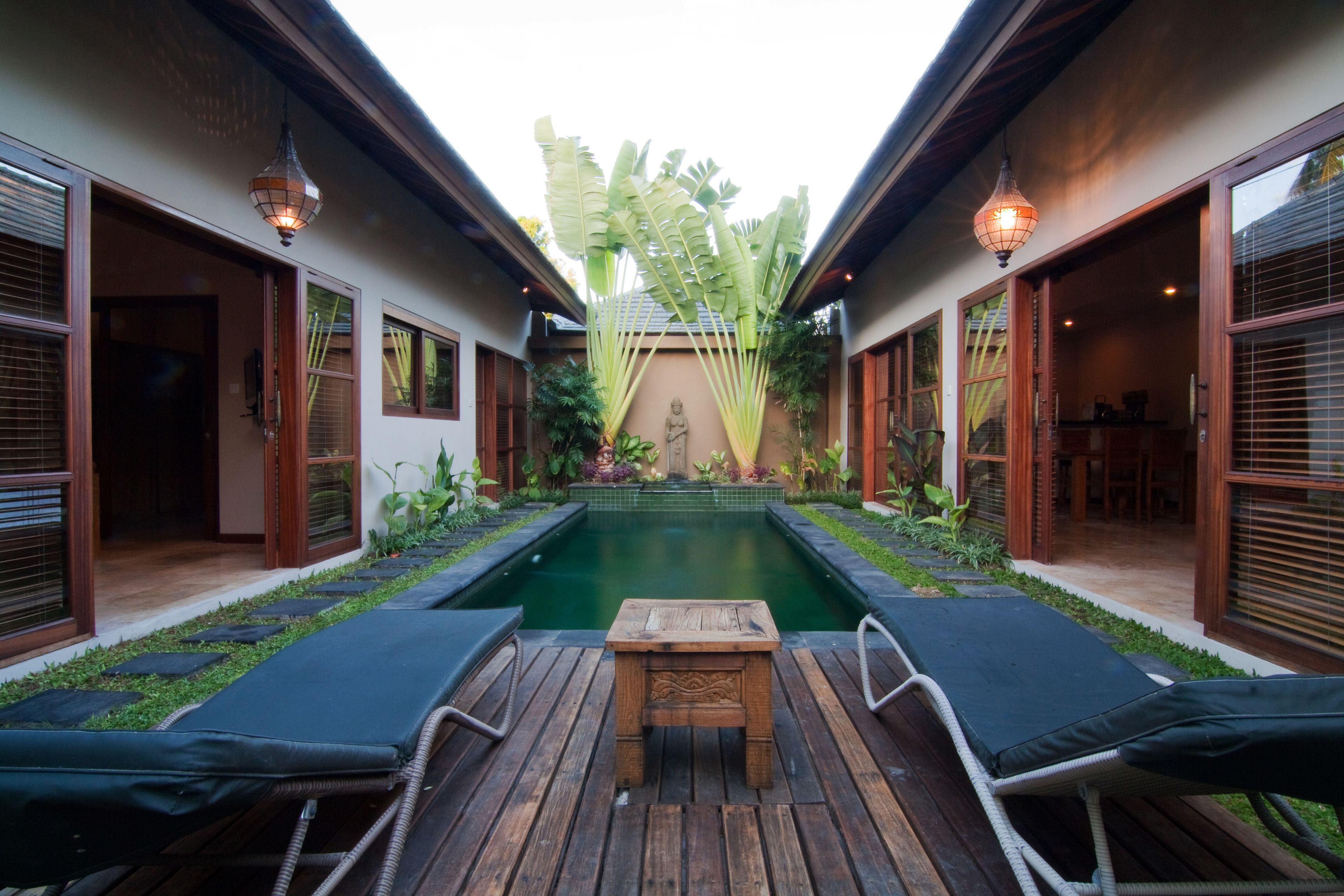 A pool area with lounge chairs and a wooden table, surrounded by plants and a statue.