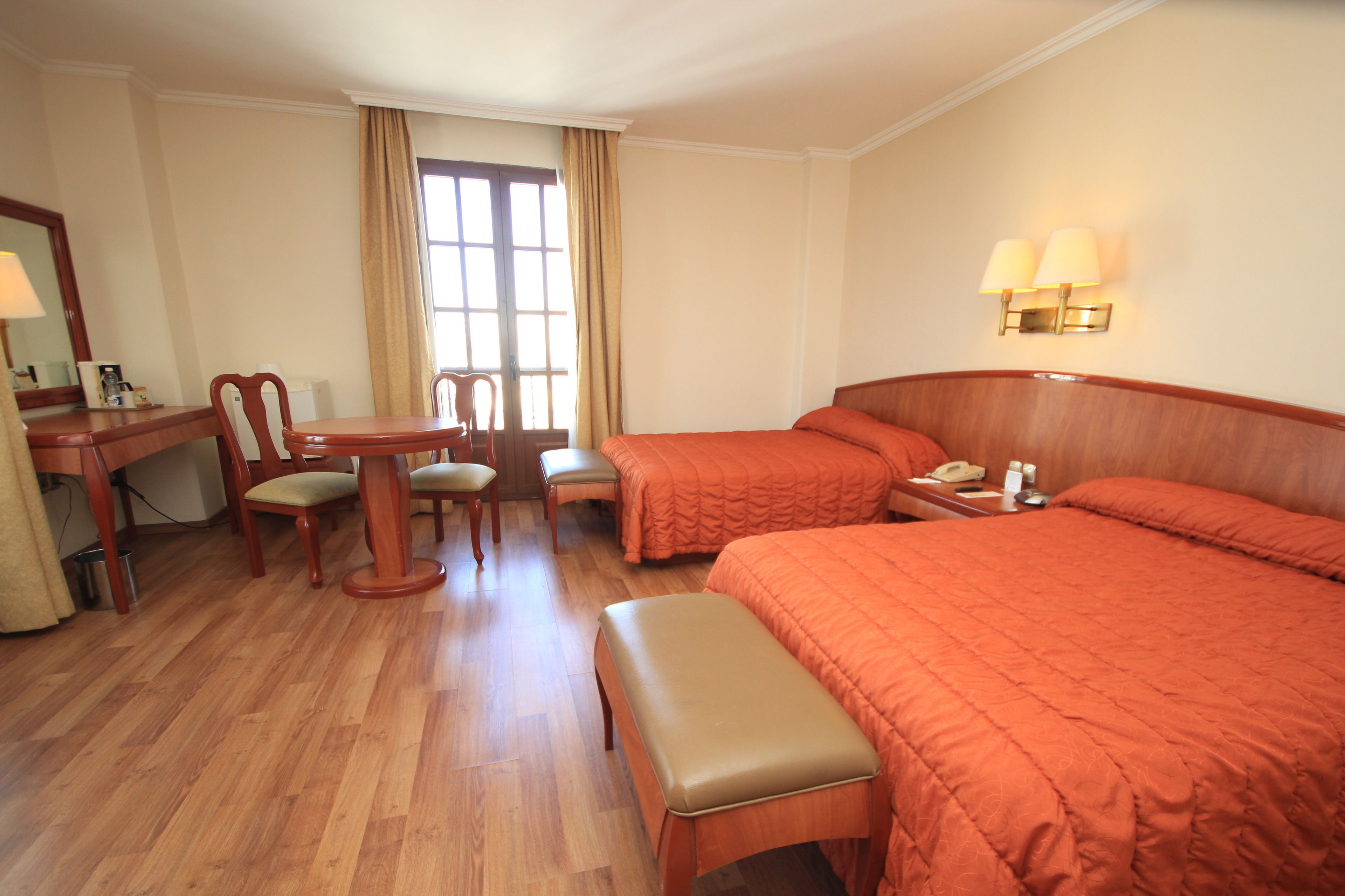 Una habitación de hotel con piso de madera, dos camas individuales con ropa de cama naranja, un escritorio de madera con una silla, una mesita con otra silla, y una ventana con cortinas.