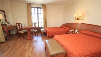 Una habitación de hotel con piso de madera, dos camas individuales con ropa de cama naranja, un escritorio de madera con una silla, una mesita con otra silla, y una ventana con cortinas.