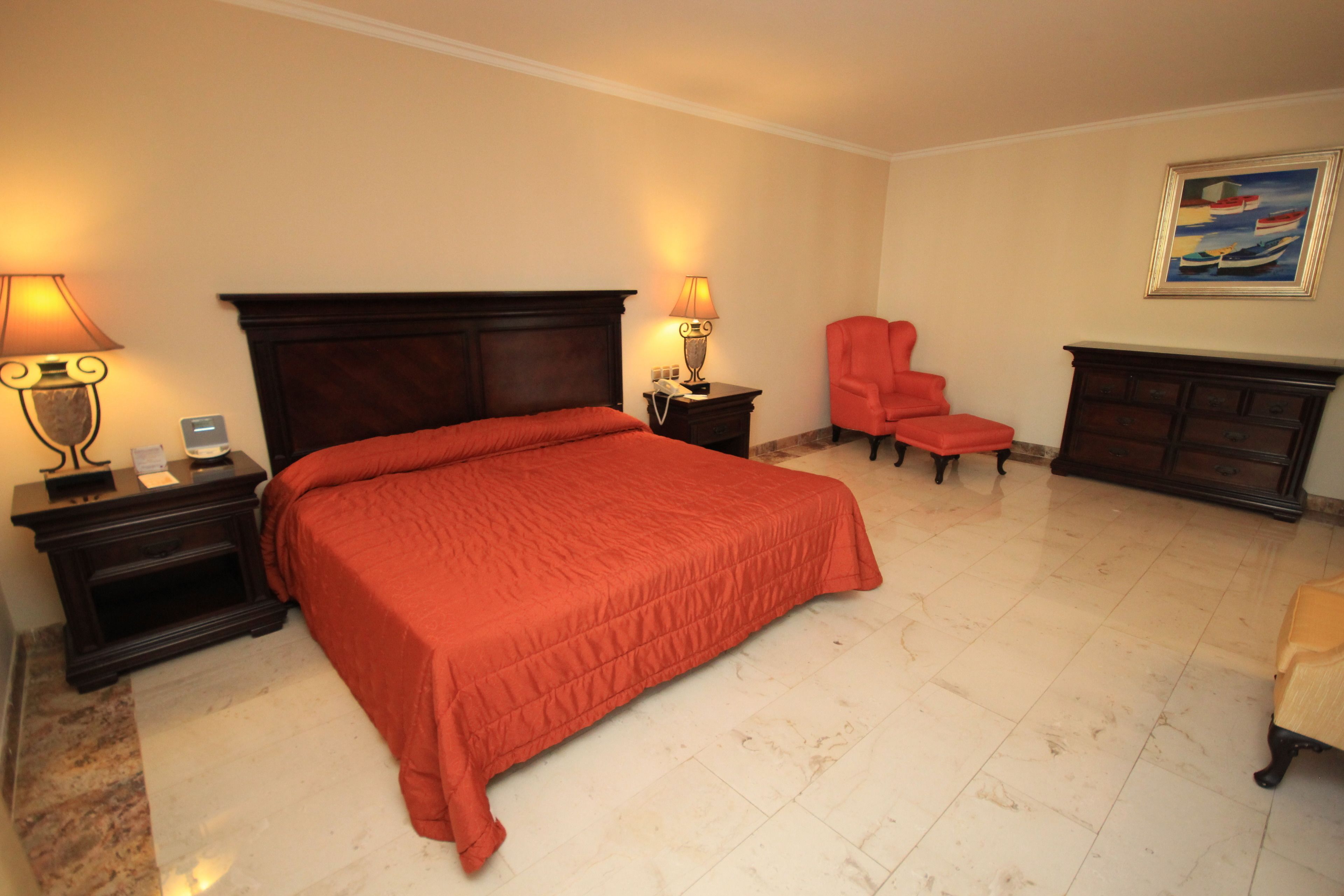 Una camera d'albergo con un letto grande, due comodini con lampade, una poltrona rossa, una cassettiera e un quadro appeso al muro.