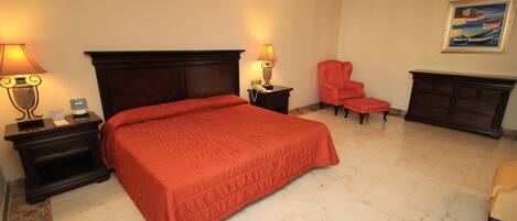 A hotel room with a large bed, two bedside tables with lamps, a red chair, a dresser, and a painting on the wall.