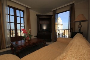 Habitación de hotel con una cama grande, un televisor sobre un mueble de madera, una mesa con un jarrón de flores y un balcón con vistas a un edificio.