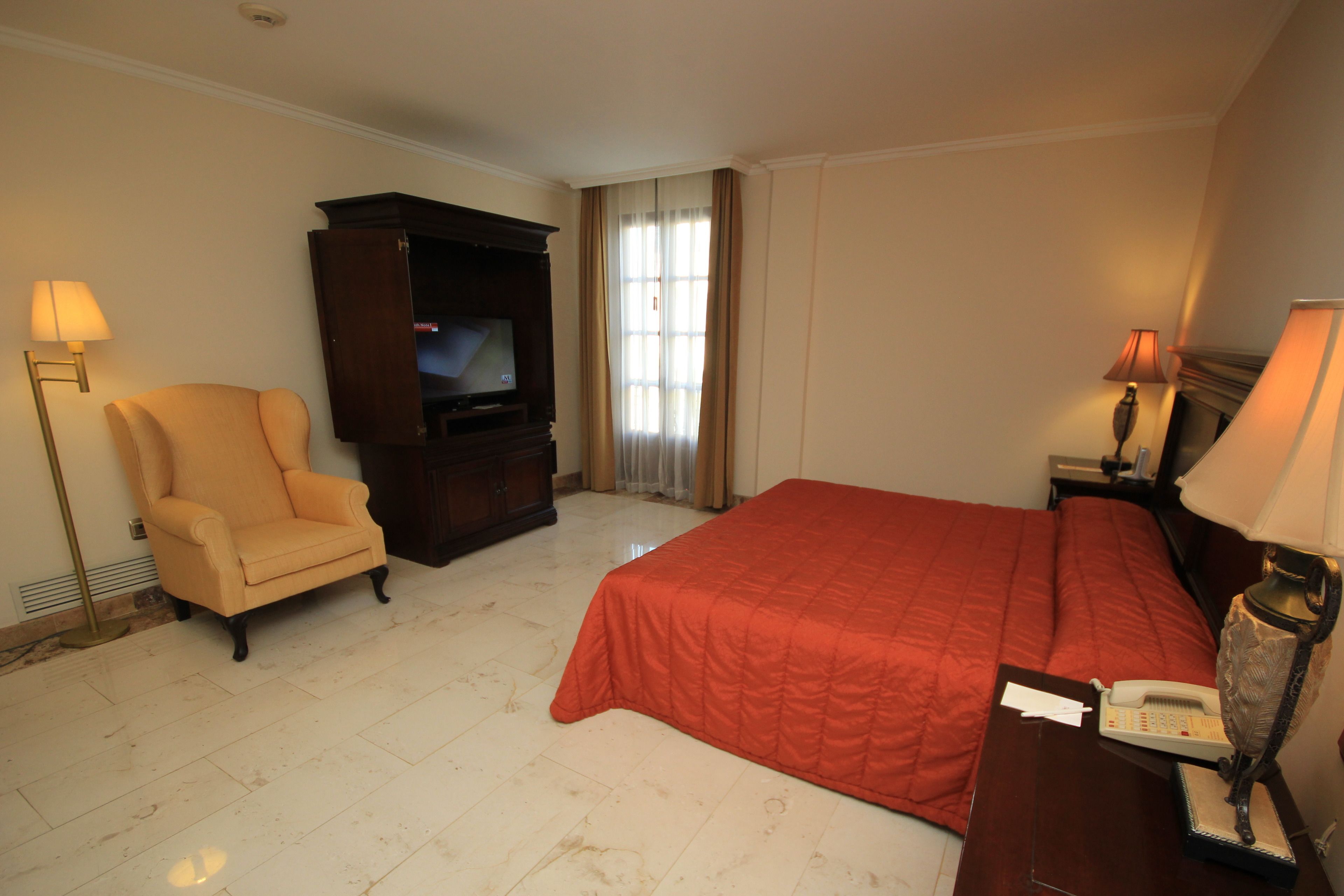 Habitación de hotel con cama, una silla, televisor y lámpara.
