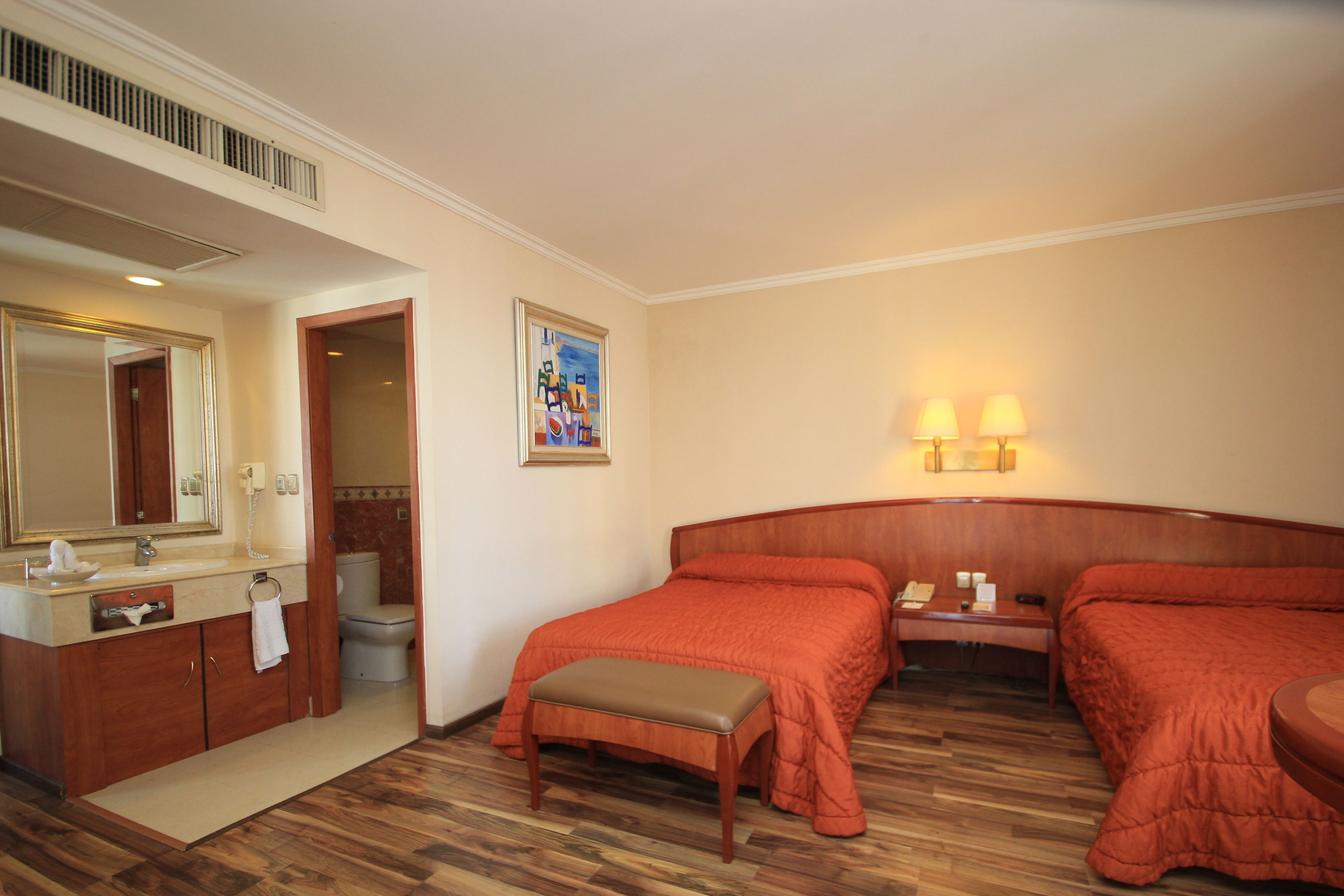 Una habitación de hotel con piso de madera, una cama con cubrecama naranja, una mesita de noche con lámpara, un baño con espejo y lavamanos, y un cuadro en la pared.