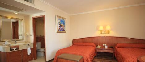 Una habitación de hotel con piso de madera, una cama con cubrecama naranja, una mesita de noche con lámpara, un baño con espejo y lavamanos, y un cuadro en la pared.