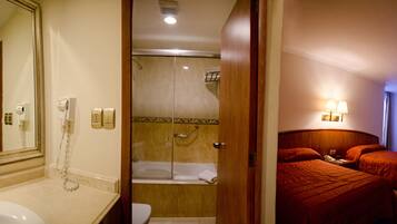 Casa de banho de hotel com lavatório, espelho e chuveiro. O quarto possui uma porta de madeira e uma cama com roupa de cama vermelha.