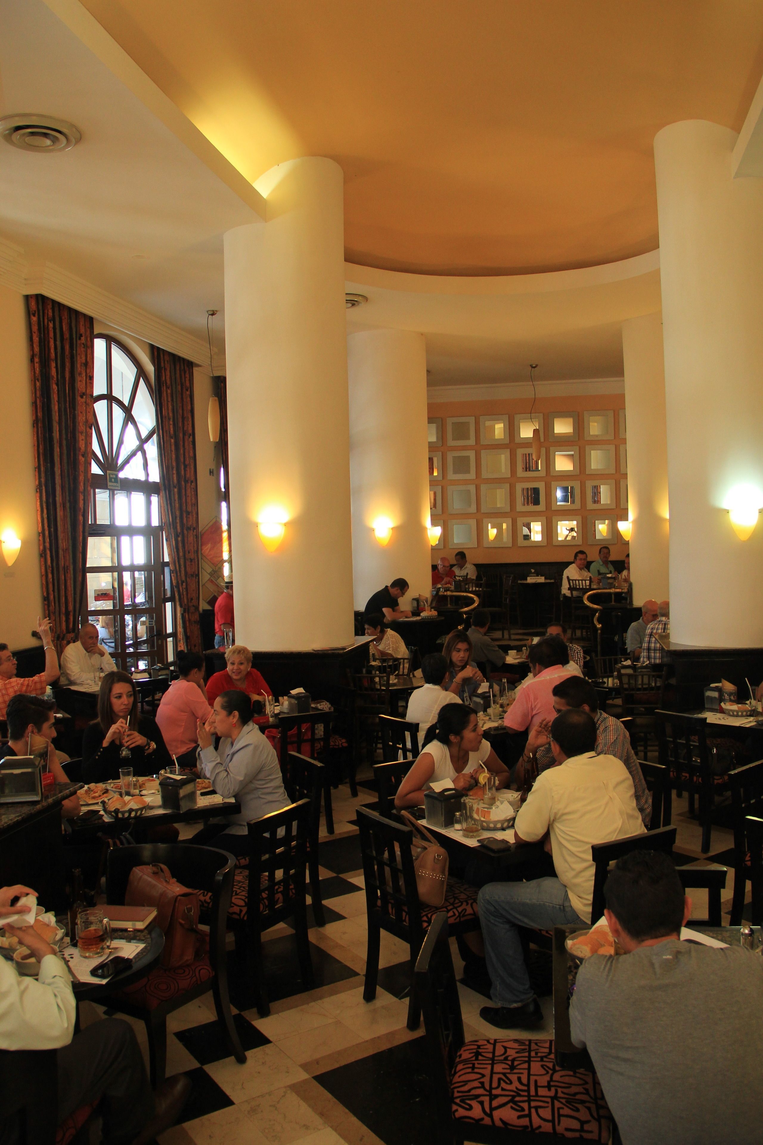 A restaurant with multiple tables and chairs, a checkered floor, and a large window with curtains.