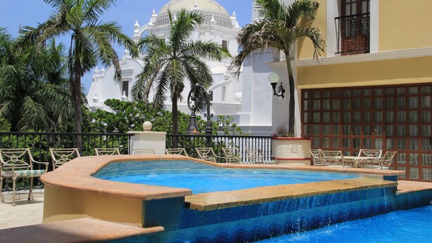 A swimming pool with a view of a white building with a dome, surrounded by palm trees and lounge chairs. - Gran Hotel Diligencias (Veracruz)