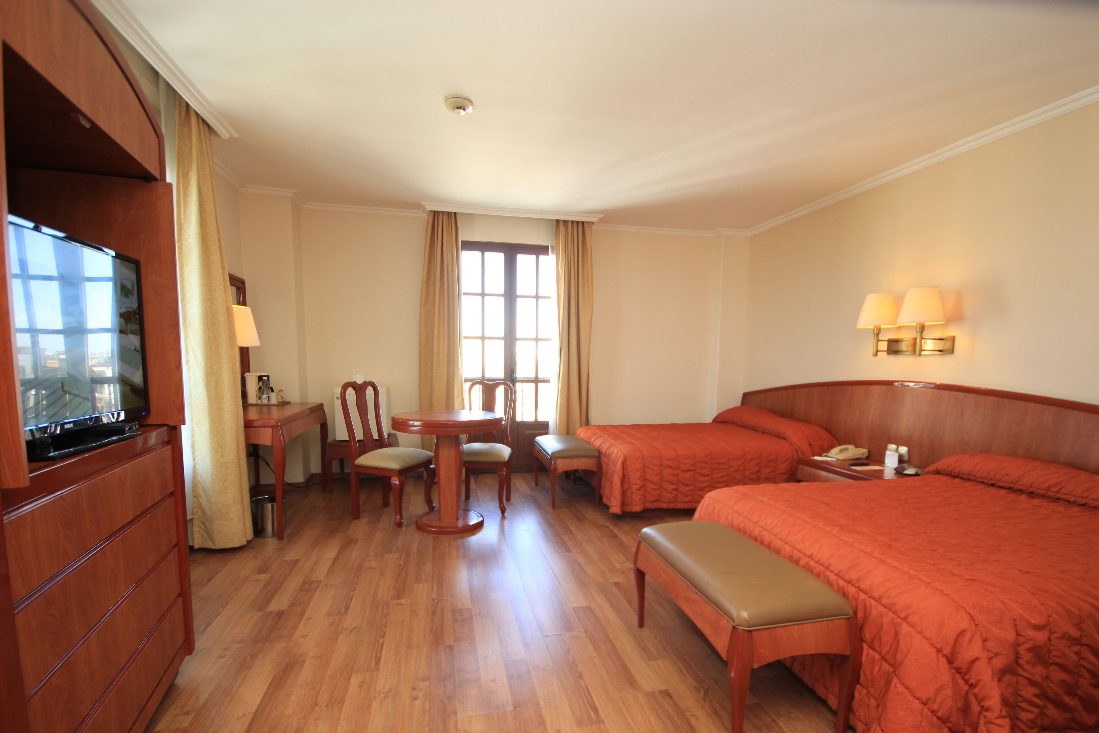 Una habitación de hotel con dos camas, un escritorio con sillas, un televisor y una ventana con cortinas.