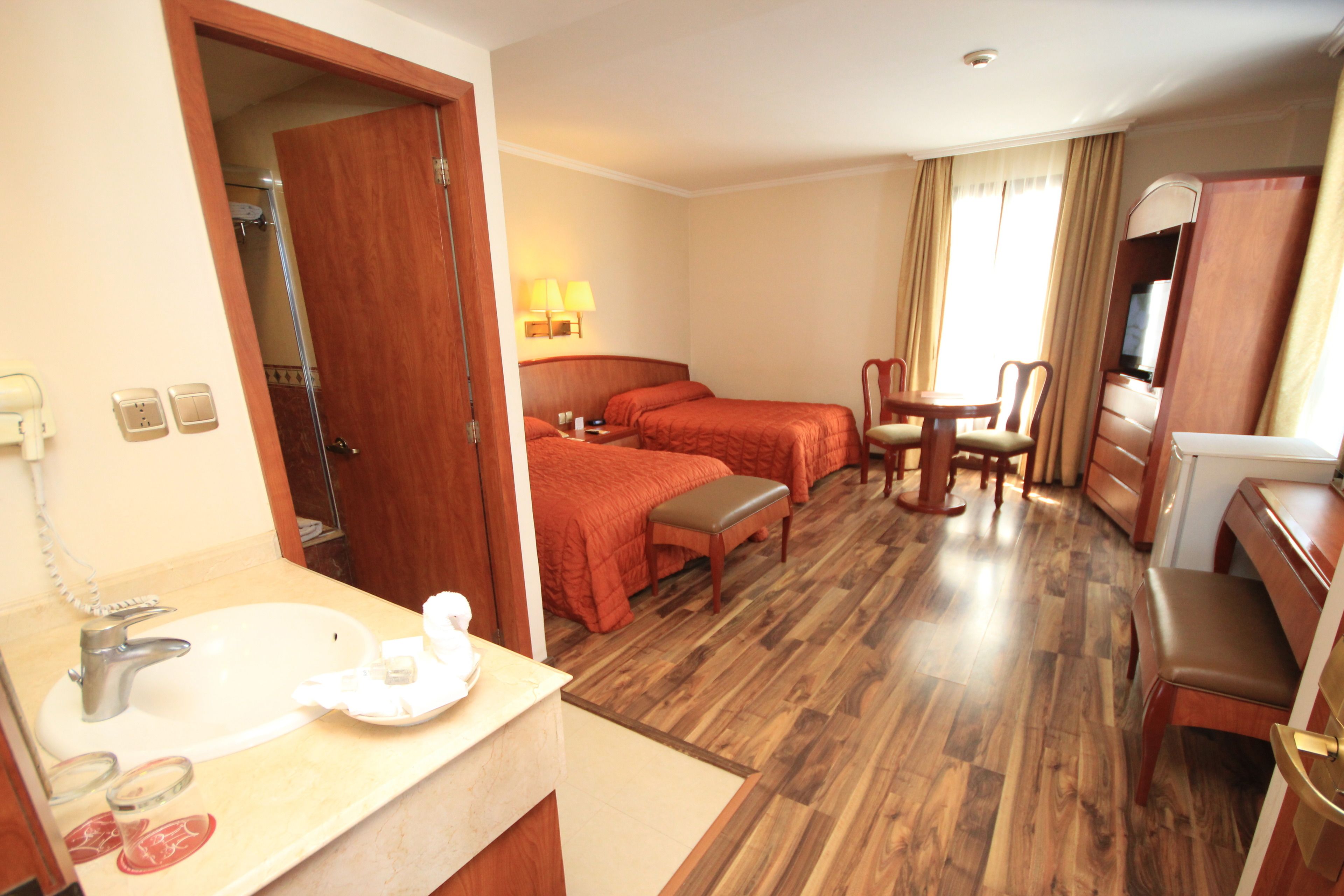 A hotel room with a wooden floor, a bed, a desk, a chair, a table, a lamp, a TV, and a bathroom with a sink and mirror.