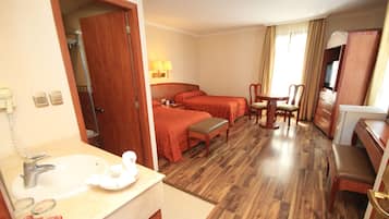 A hotel room with a wooden floor, a bed, a desk, a chair, a table, a lamp, a TV, and a bathroom with a sink and mirror.