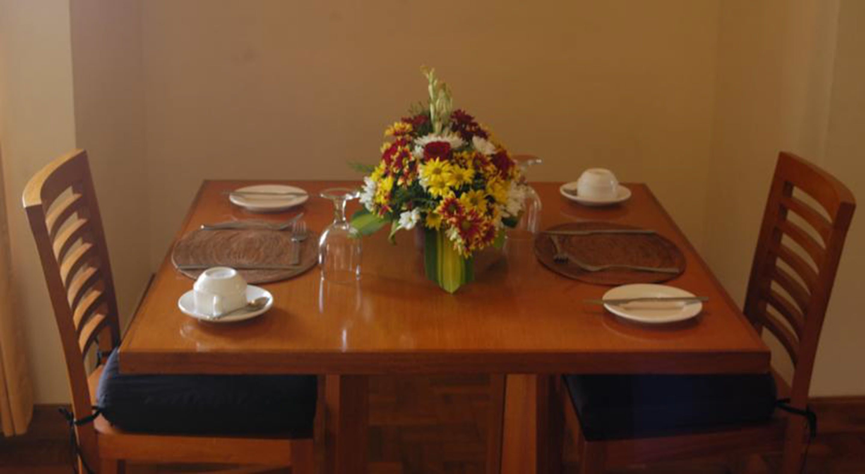 A wooden dining table with a vase of flowers, plates, cutlery, and a cup.