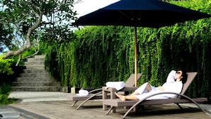 Uma pessoa relaxando em uma espreguiçadeira sob um grande guarda-sol, com uma escadaria e vegetação exuberante ao fundo.