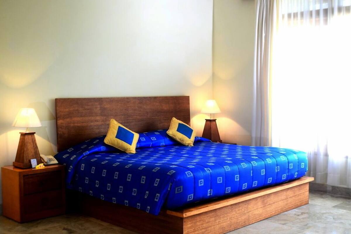 Een net opgemaakt bed met een blauw geborreld deken en twee kussens, aan weerszijden voorzien van nachtkastjes met lampen, in een kamer met een groot raam en gordijnen.
