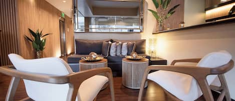 Un salon moderne avec un canapé, deux fauteuils, une table basse et une table d’appoint. Un grand miroir est placé au-dessus du canapé, et une plante en pot se trouve sur la table d’appoint.