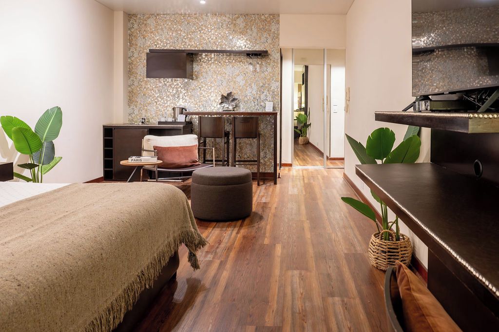 Una habitación de hotel moderna con una cama, zona de estar con mesa y sillas, kitchenette y un ventanal grande.
