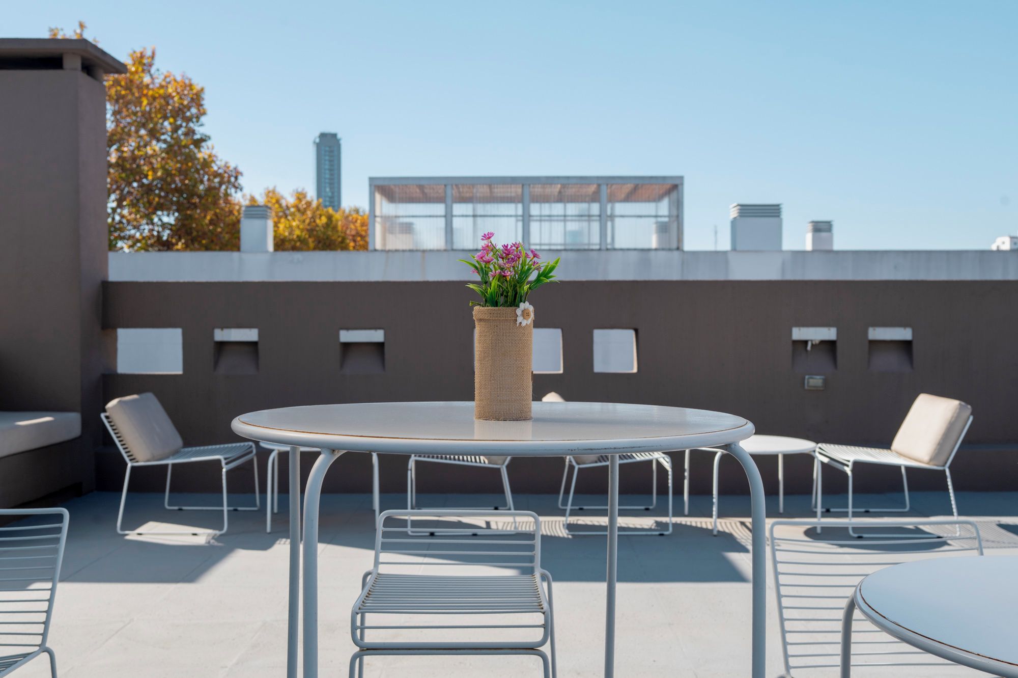 Un área de estar al aire libre con mesas redondas y sillas metálicas, un jarrón tejido con flores y un edificio moderno al fondo.