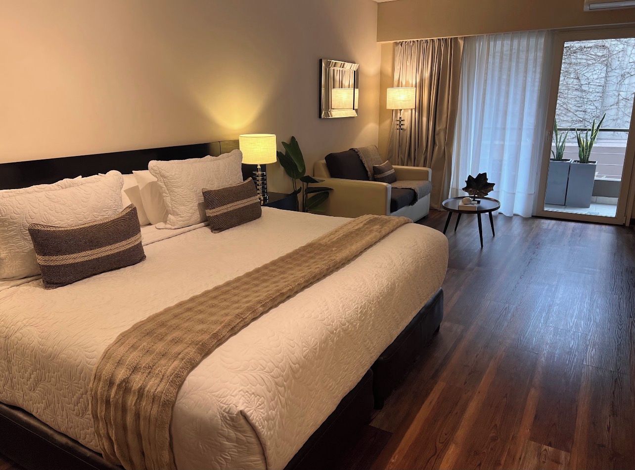 Habitación de hotel con una cama grande, un área de descanso con una silla y una mesita, y vistas a través de una ventana.