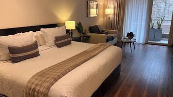 Habitación de hotel con una cama grande, un área de descanso con una silla y una mesita, y vistas a través de una ventana.