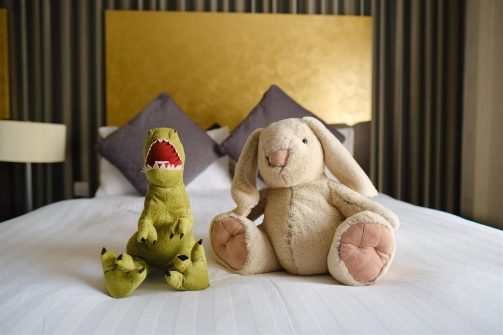 A bed with two plush toys, a green dinosaur and a white rabbit, in a room with a yellow headboard and striped wallpaper.