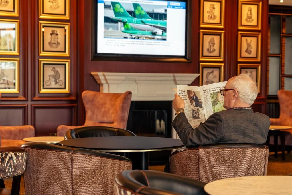 Un hombre leyendo un periódico en una habitación con cuadros en la pared y un televisor mostrando un avión.