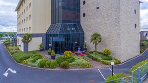 A modern hotel with a glass entrance, a stone tower, and a landscaped garden with neatly trimmed bushes and a palm tree. - Talbot Hotel Carlow (Carlow)
