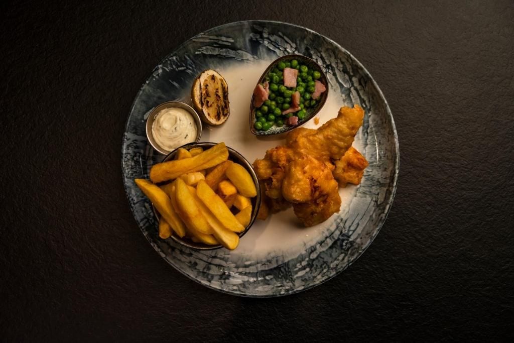 A plate with fish and chips, peas, and a small bowl of sauce.