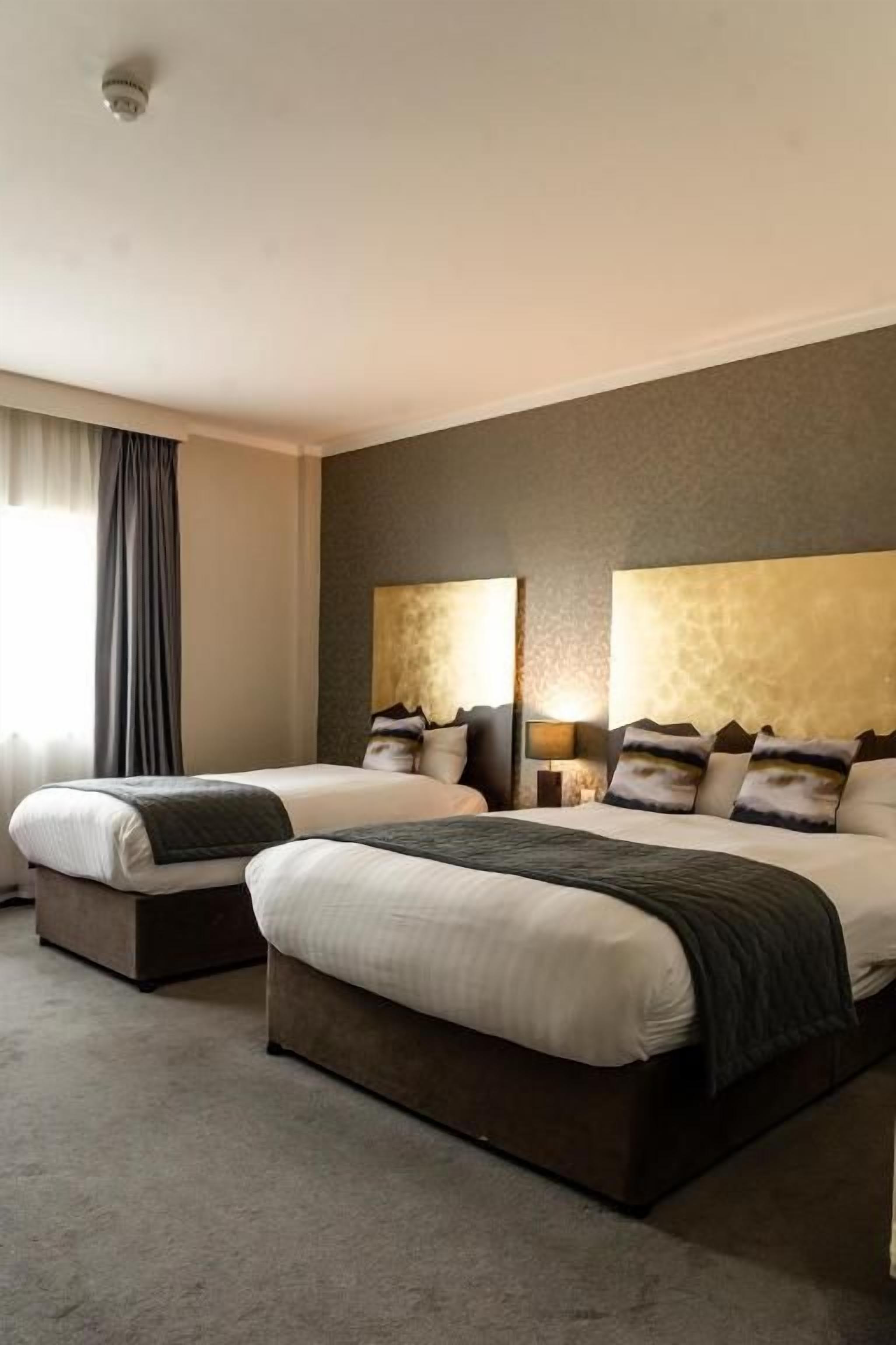 A hotel room with two beds, a large window with curtains, and a wall-mounted lamp.