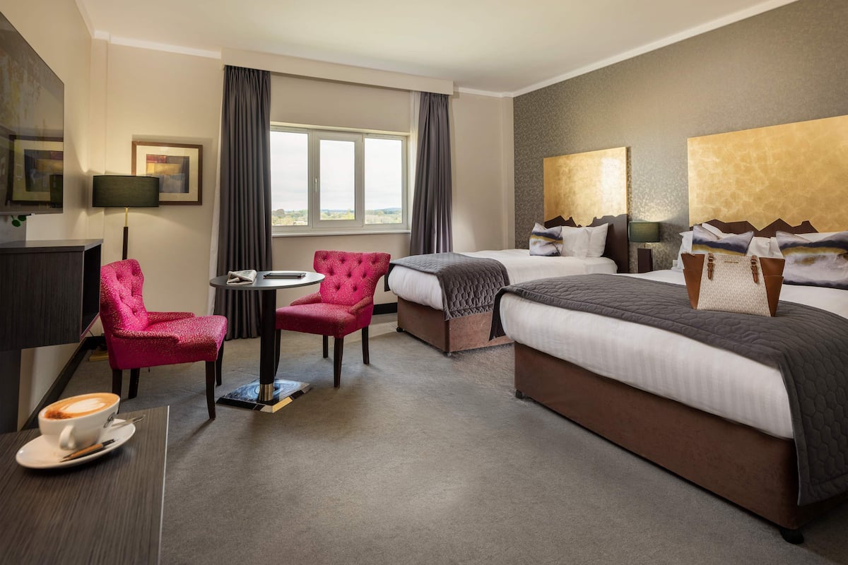 A hotel room with a large bed, two smaller beds, a desk with a chair, a coffee table, and a view of the city through the window.