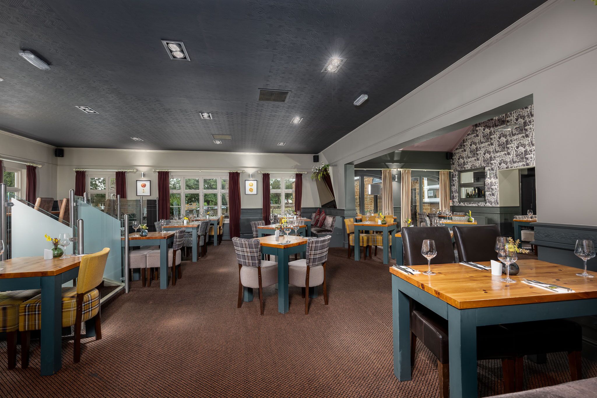 A restaurant with multiple dining tables and chairs, a patterned ceiling, and large windows with curtains.