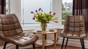 Quarto com duas poltronas de couro marrom, uma pequena mesa de madeira com um vaso de tulipas roxas e vista para o exterior pela janela.