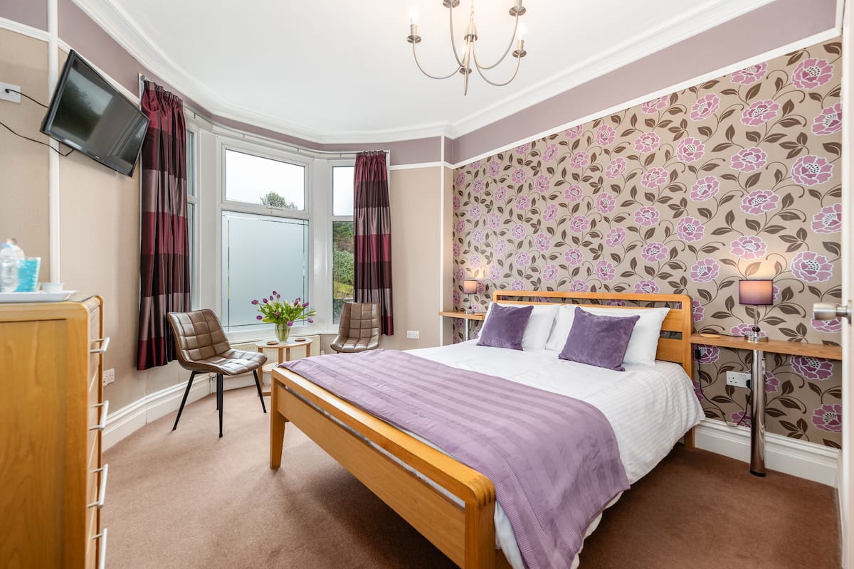 A bedroom with a bed, a wooden bench, a chair, a TV mounted on the wall, and a window with curtains.