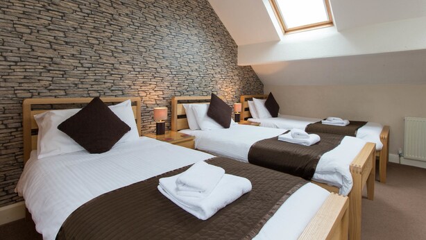 Quarto de hotel com três camas, parede de pedra, cabeceira de madeira e um claraboia.