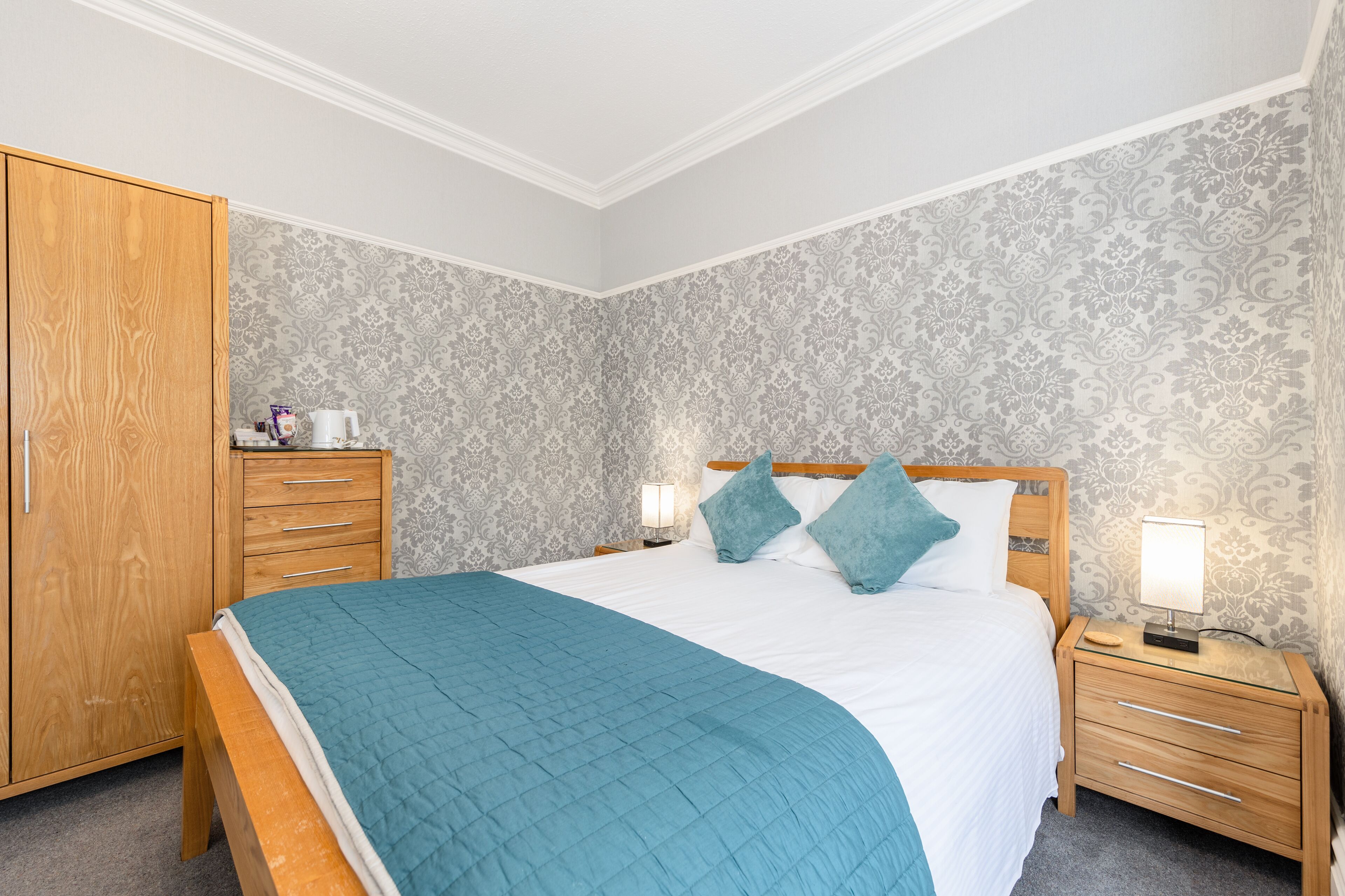A bedroom with a bed, a wooden wardrobe, a nightstand with a lamp, and a patterned wallpaper.