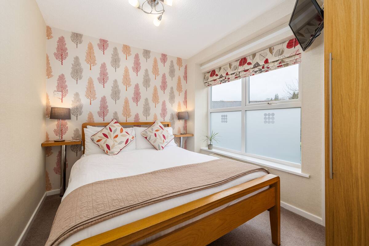 A bedroom with a bed, bedside lamps, a window with a view, and a TV mounted on the wall.