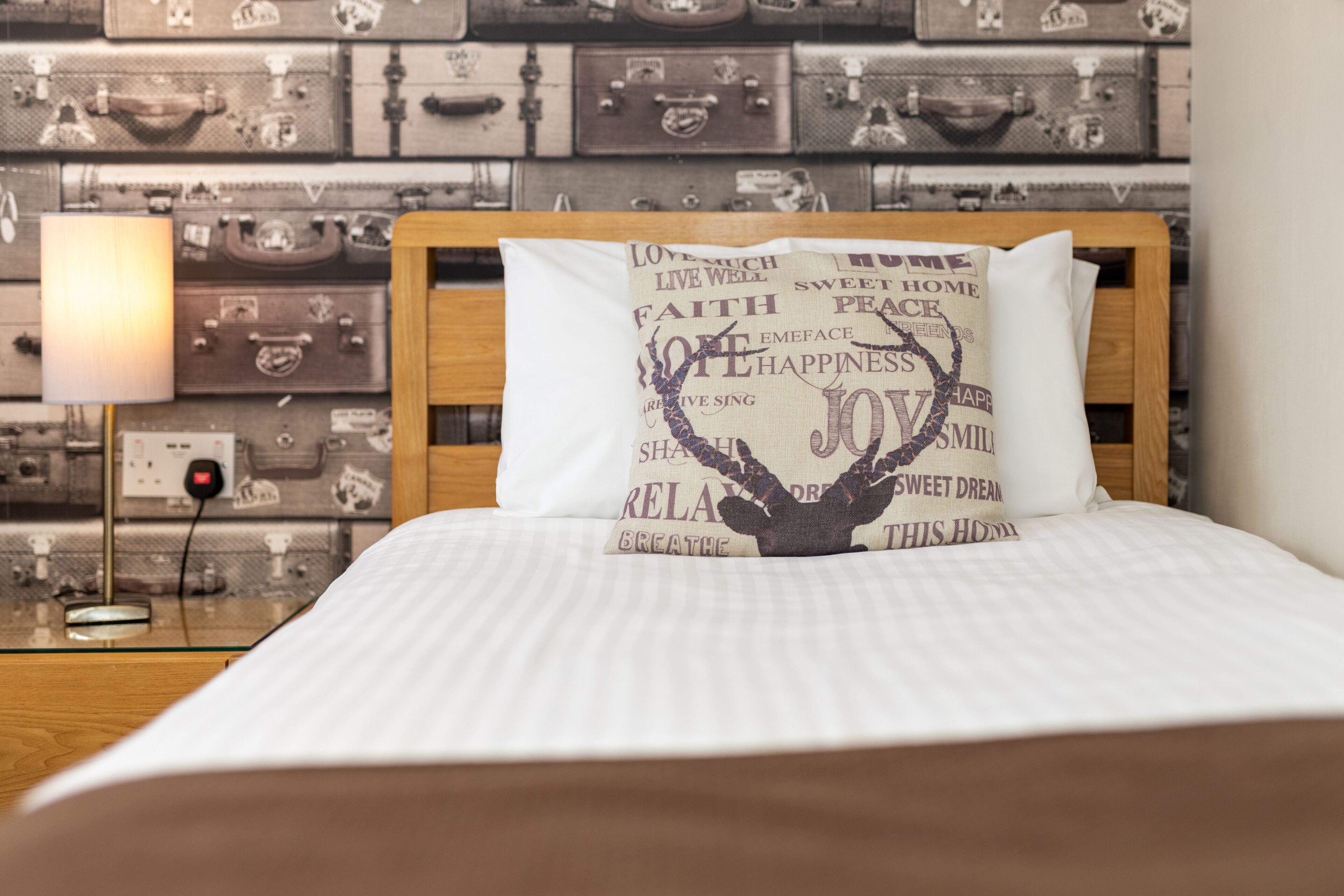 Un letto rifatto con cura, con un cuscino decorato da parole motivazionali, una lampada da comodino e una parete di valigie vintage.