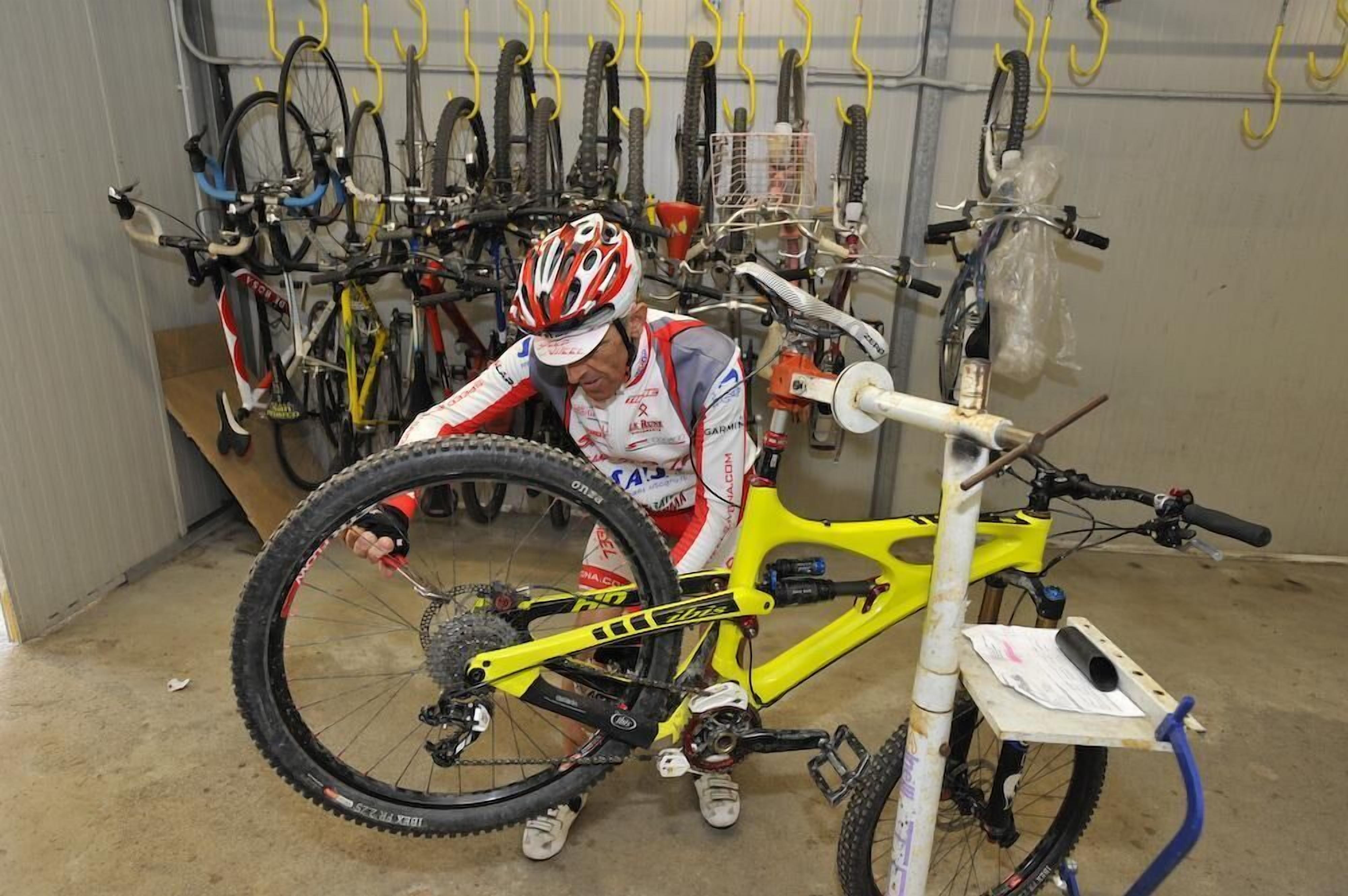 Une personne est en train de travailler sur un vélo de montagne jaune dans un garage où se trouvent plusieurs vélos et des pneus accrochés au mur.