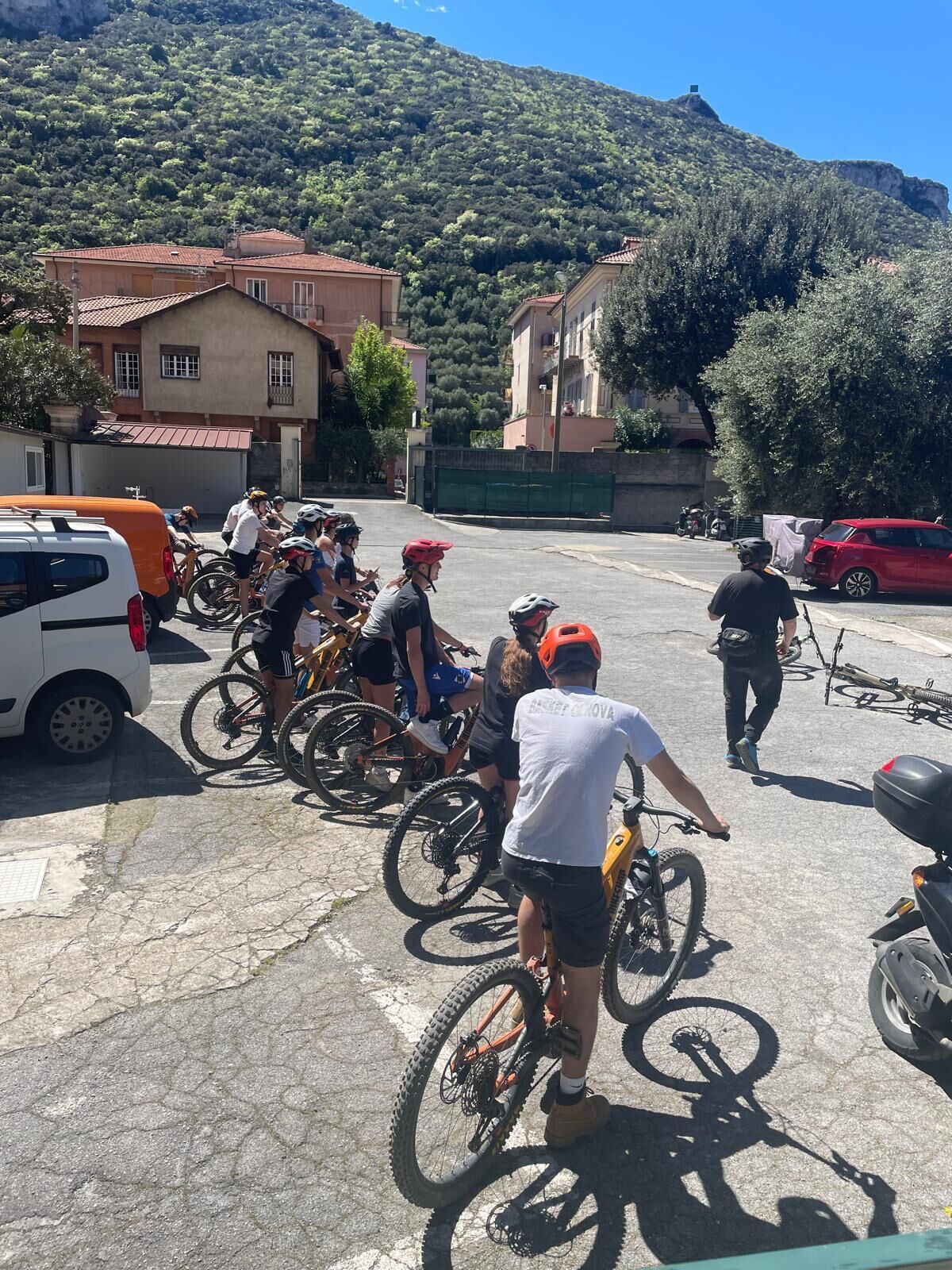 A group of cyclists gathered in a parking lot with buildings and a mountain in the background.