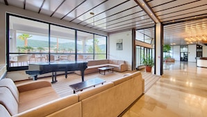 A spacious living area with a sofa, a piano, and a dining table. Large windows offer a view of the outdoors. - Hotel Issa (Vis)