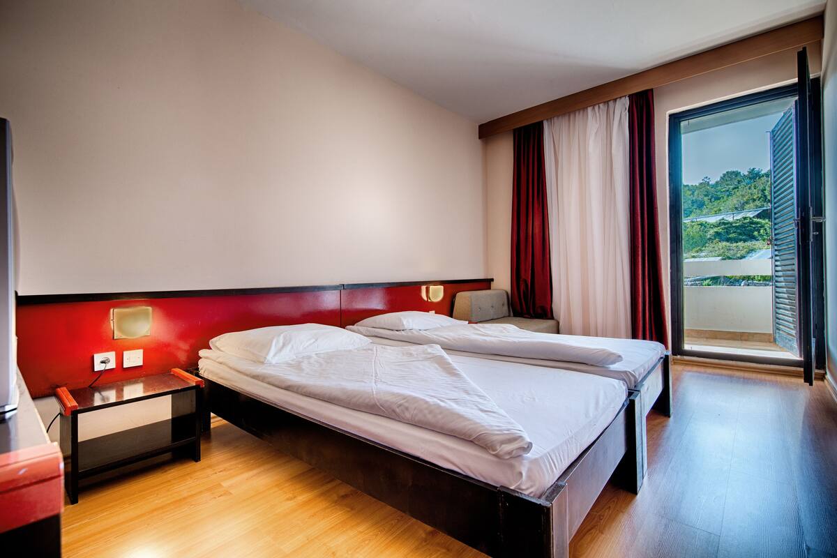 A hotel room with two beds, a red accent wall, a nightstand, and a view of greenery through the window.