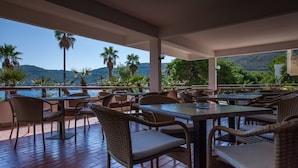 An outdoor dining area with wicker furniture and a view of hills and palm trees. - Hotel Issa (Vis)