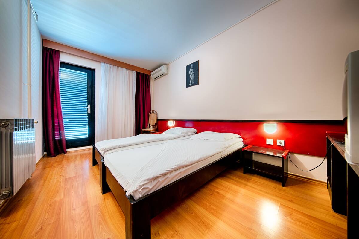 A bedroom with a wooden floor, a bed, a nightstand, a red headboard, a wall-mounted air conditioner, and a window with curtains.