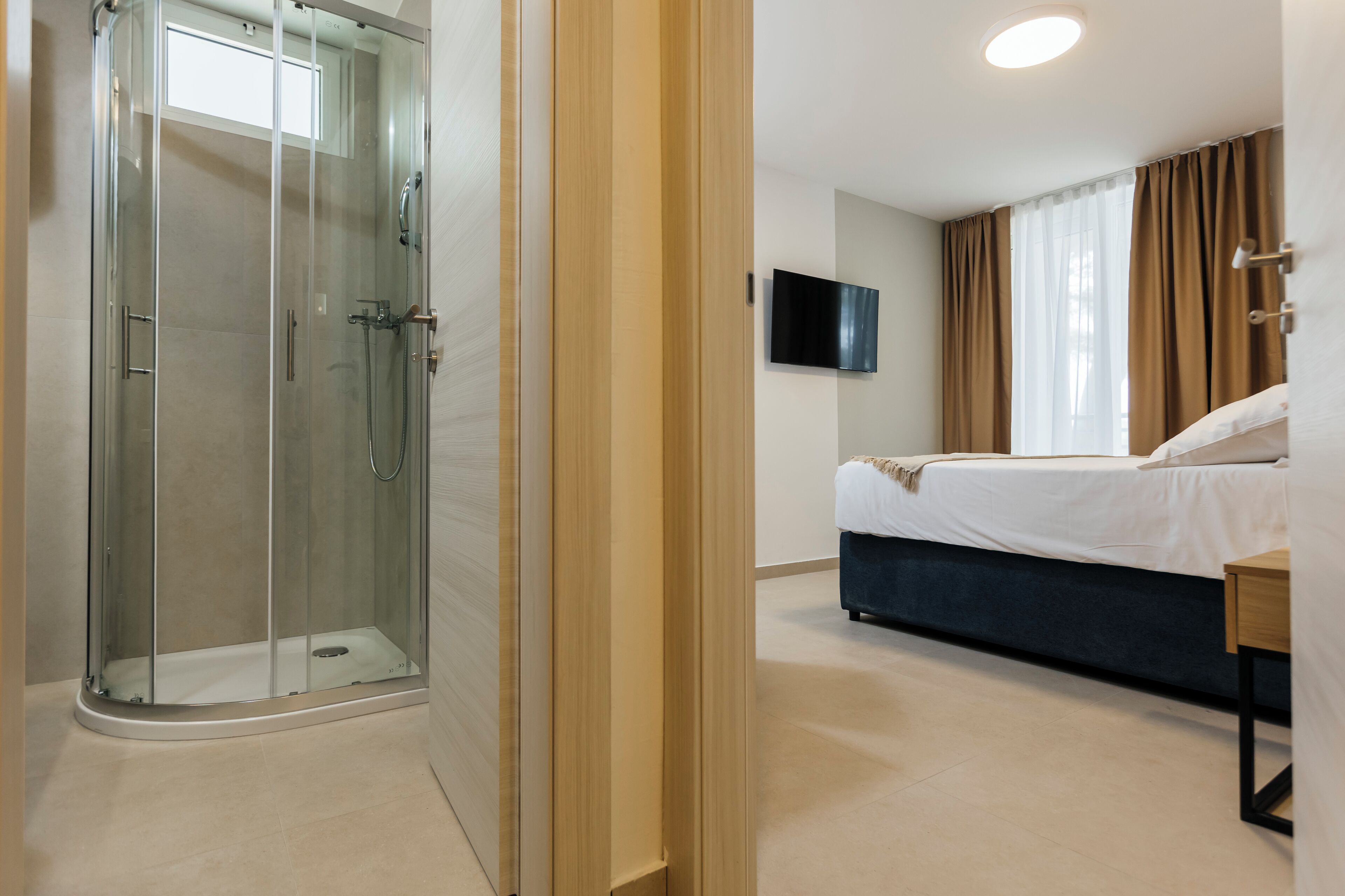 A hotel room with a glass-enclosed shower, a bed with a dark headboard, a flat-screen TV mounted on the wall, and a window with curtains.